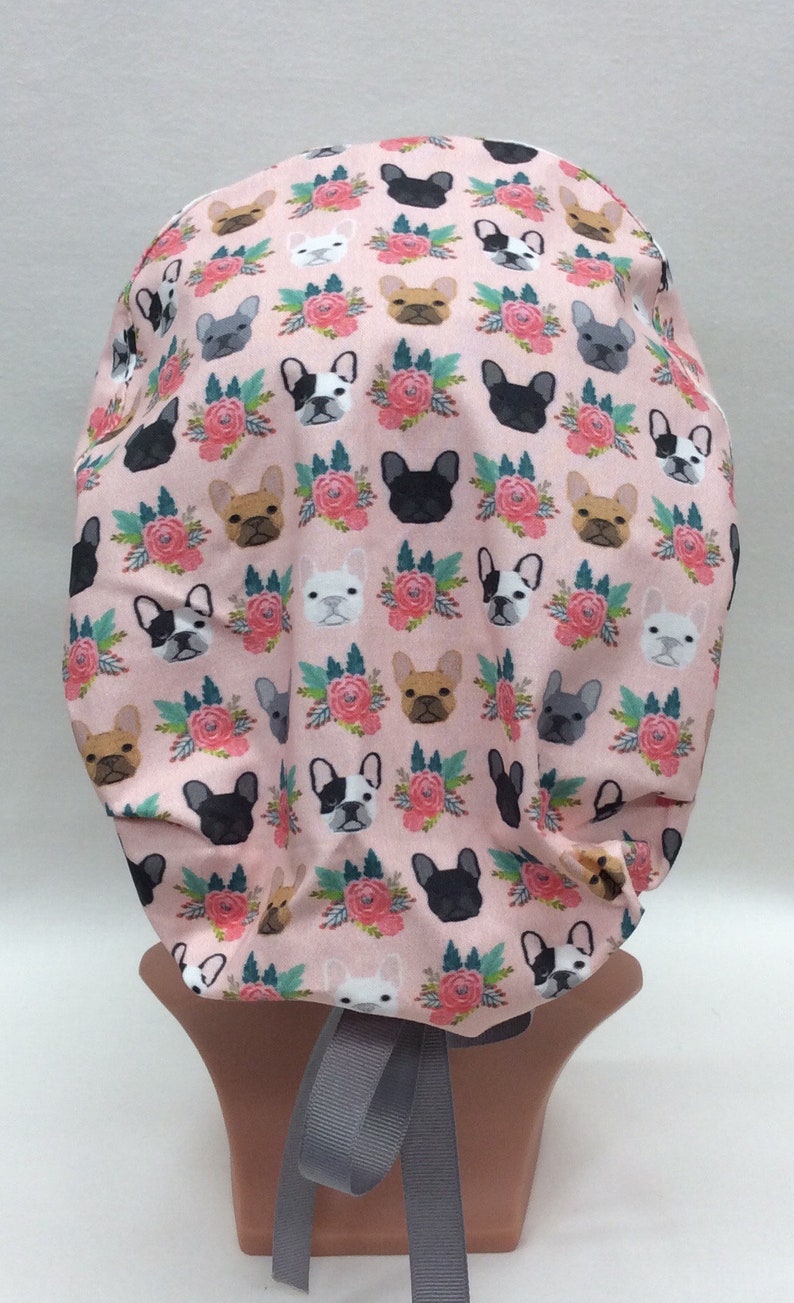 May include: A pink surgical scrub hat with a pattern of French Bulldogs and floral bouquets. The hat has a gray ribbon tie.