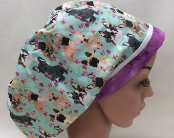May include: A light blue surgical scrub cap featuring a repeating pattern of French Bulldogs in various colors. The cap is trimmed with a purple band and white piping. The design includes pink floral accents.