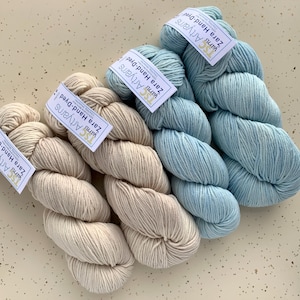 May include: Close-up of hand-dyed yarn skeins in light blue and cream colors. Each skein has a tag that reads "Zara Hand-Dyed" and "Artyarns". The yarn is arranged in a visually appealing pattern, showcasing the texture and color variations.