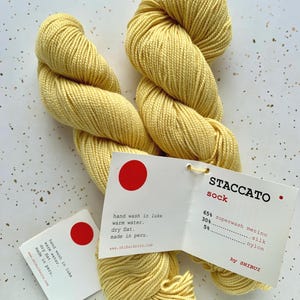 May include: Two skeins of pale yellow Staccato sock yarn with attached tags. The tags include washing instructions and the yarn's composition: 65% superwash merino, 30% silk, and 5% nylon. The yarn is made in Peru.
