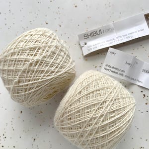 May include: Two balls of ivory Shibui Cima yarn, composed of 70% super baby alpaca and 30% fine merino wool. Each 1.76 oz ball contains 328 yards. The labels indicate the color is ivory, hand wash only, and the product is made in Peru.