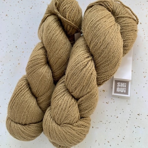 May include: Two skeins of tan-colored yarn are displayed against a speckled white background. The yarn is thick and twisted, with a soft, textured appearance. A white tag with the text "SHIBUI" is attached to one skein.