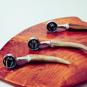 Puede incluir: Tres cucharas para helado con cuencos de metal pulido y mangos de asta descansan sobre una tabla de cortar de madera marrón rojizo. Las cucharas están dispuestas en fila, mostrando su diseño único y artesanía. La tabla tiene un acabado liso y pulido.