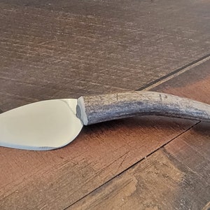 Op de afbeelding: Een kaasmes met een gepolijst, zilverkleurig lemmet en een handvat van een getextureerd, bruin gewei. Het mes heeft een gebogen lemmet en is ontworpen voor het snijden van zachte kazen. Het mes is ongeveer 18 cm lang.