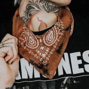 May include: A person wearing a black t-shirt with the word "IOWA" printed on it. They are wearing a brown bandana with a white paisley pattern around their neck. The bandana has a tie-dye effect.