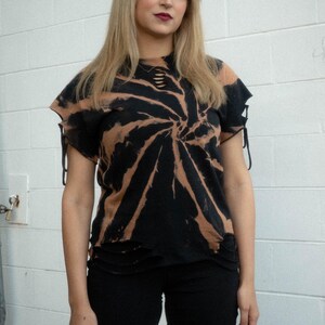 May include: A black and tan spiral tie-dye t-shirt featuring distressed cutouts at the neckline and hem, along with side sleeve ties. The shirt is paired with solid black fitted shorts that reach above the knee, creating a coordinated outfit.