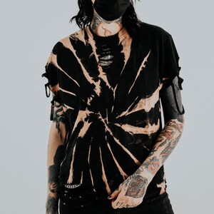 K&ouml;nnte beinhalten: Ein schwarzes Kurzarm-T-Shirt mit einem gebleichten Spiral-Tie-Dye-Muster, einem abgenutzten Halsausschnitt mit vertikalen Schlitzen und kleinen seitlichen Bindeb&auml;ndern an den &Auml;rmeln. Das Shirt wird mit einer schwarzen, abgenutzten Jeans mit gro&szlig;en Knierissen kombiniert.