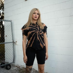 K&ouml;nnte beinhalten: Ein schwarz-beiges Batik-T-Shirt mit Spiralmuster, Distressed-Details an Ausschnitt und Saum sowie kurzen &Auml;rmeln mit seitlichen Bindeb&auml;ndern, kombiniert mit einer einfarbigen, eng anliegenden schwarzen Shorts bis &uuml;ber das Knie.