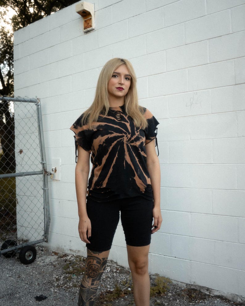 K&ouml;nnte beinhalten: Ein schwarz-beiges Batik-T-Shirt mit Spiralmuster, Distressed-Details an Ausschnitt und Saum sowie kurzen &Auml;rmeln mit seitlichen Bindeb&auml;ndern, kombiniert mit einer einfarbigen, eng anliegenden schwarzen Shorts bis &uuml;ber das Knie.