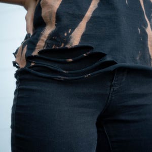 K&ouml;nnte beinhalten: Ein schwarzes T-Shirt mit gebleichtem Batikmuster, diagonalen Streifen und horizontalen, fransigen Einschnitten am unteren Saum, kombiniert mit einer schlichten schwarzen Jeans. Das T-Shirt hat einen unges&auml;umten Rand und dunkle Farben.