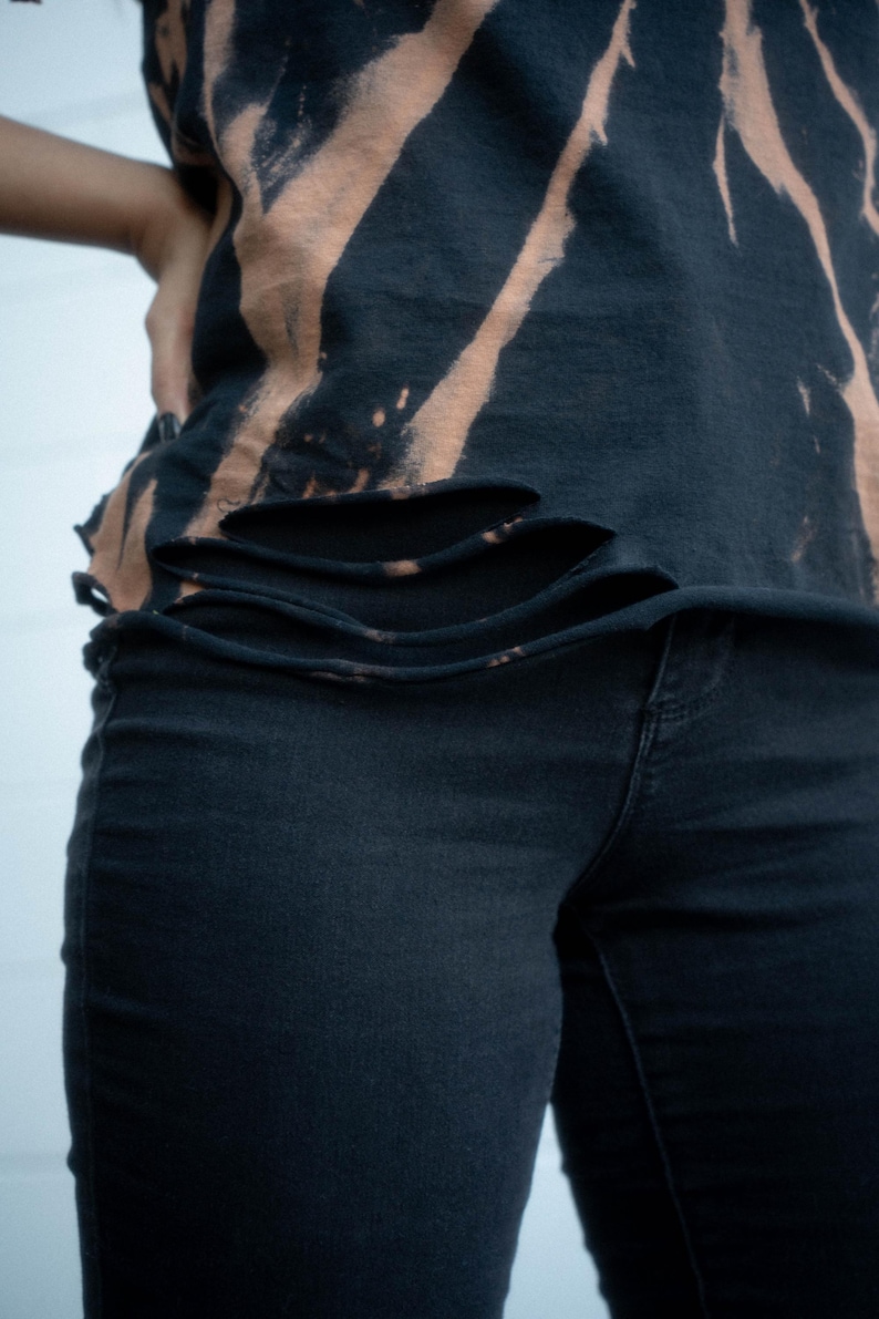 K&ouml;nnte beinhalten: Ein schwarzes T-Shirt mit gebleichtem Batikmuster, diagonalen Streifen und horizontalen, fransigen Einschnitten am unteren Saum, kombiniert mit einer schlichten schwarzen Jeans. Das T-Shirt hat einen unges&auml;umten Rand und dunkle Farben.