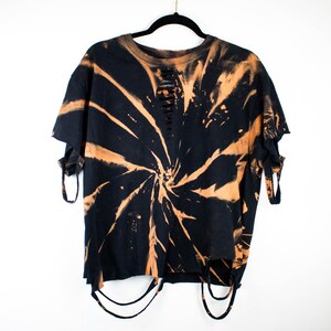 K&ouml;nnte beinhalten: Ein schwarzes Kurzarm-T-Shirt mit einem spiralf&ouml;rmigen, orangefarbenen Bleichmuster. Das Kleidungsst&uuml;ck weist Distressed-Details mit horizontalen Schlitzen am Halsausschnitt, ausgefransten &Auml;rmelkanten und d&uuml;nnen Stoffstreifen am Saum auf.