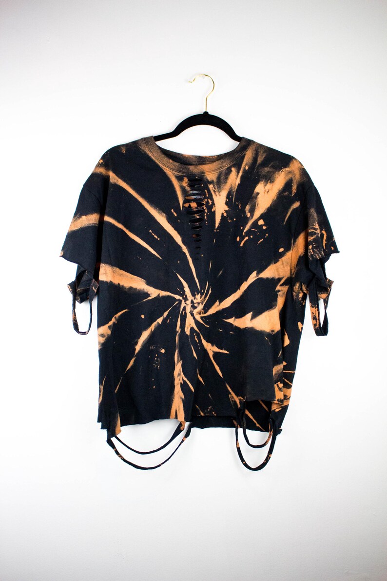 K&ouml;nnte beinhalten: Ein schwarzes Kurzarm-T-Shirt mit einem spiralf&ouml;rmigen, orangefarbenen Bleichmuster. Das Kleidungsst&uuml;ck weist Distressed-Details mit horizontalen Schlitzen am Halsausschnitt, ausgefransten &Auml;rmelkanten und d&uuml;nnen Stoffstreifen am Saum auf.