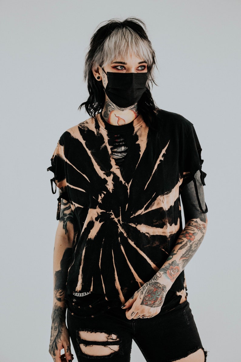 K&ouml;nnte beinhalten: Ein schwarzes Kurzarm-T-Shirt mit einem gebleichten Spiral-Tie-Dye-Muster, einem abgenutzten Halsausschnitt mit vertikalen Schlitzen und kleinen seitlichen Bindeb&auml;ndern an den &Auml;rmeln. Das Shirt wird mit einer schwarzen, abgenutzten Jeans mit gro&szlig;en Knierissen kombiniert.