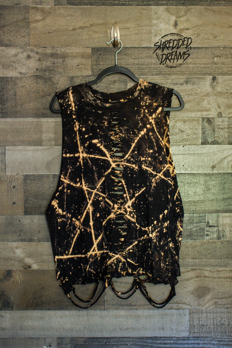 May include: Black sleeveless shirt with bleach splattered design and distressed detailing. The shirt has a round neckline and is hanging on a black hanger. The text "Shredded Dreams" is visible in the top right corner of the image.