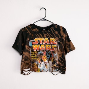 May include: A black and brown acid-wash crop top t-shirt featuring a Star Wars comic book graphic on the front. The design includes the Star Wars logo, characters, and the text A long time ago in a galaxy far away. The hem features distressed cutouts.