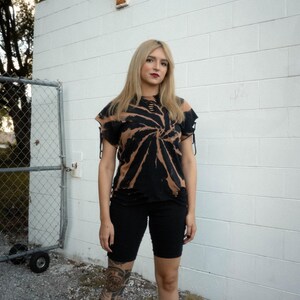 K&ouml;nnte beinhalten: Ein kurz&auml;rmeliges schwarzes T-Shirt mit gebleichtem Spiral-Tie-Dye-Muster, abgenutzten Ausschnitten am Halsausschnitt und Saum sowie seitlichen &Auml;rmelb&auml;ndern, kombiniert mit einer schwarzen, eng anliegenden Shorts, die &uuml;ber dem Knie endet.