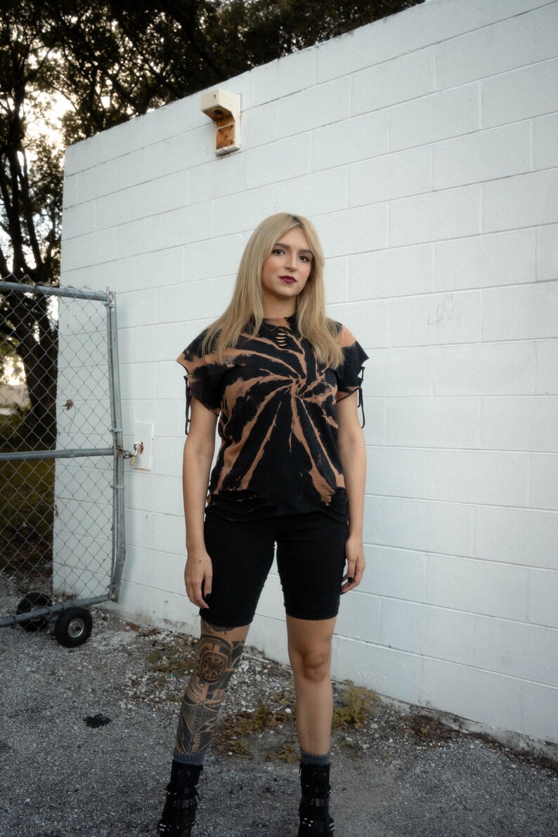 K&ouml;nnte beinhalten: Ein kurz&auml;rmeliges schwarzes T-Shirt mit gebleichtem Spiral-Tie-Dye-Muster, abgenutzten Ausschnitten am Halsausschnitt und Saum sowie seitlichen &Auml;rmelb&auml;ndern, kombiniert mit einer schwarzen, eng anliegenden Shorts, die &uuml;ber dem Knie endet.