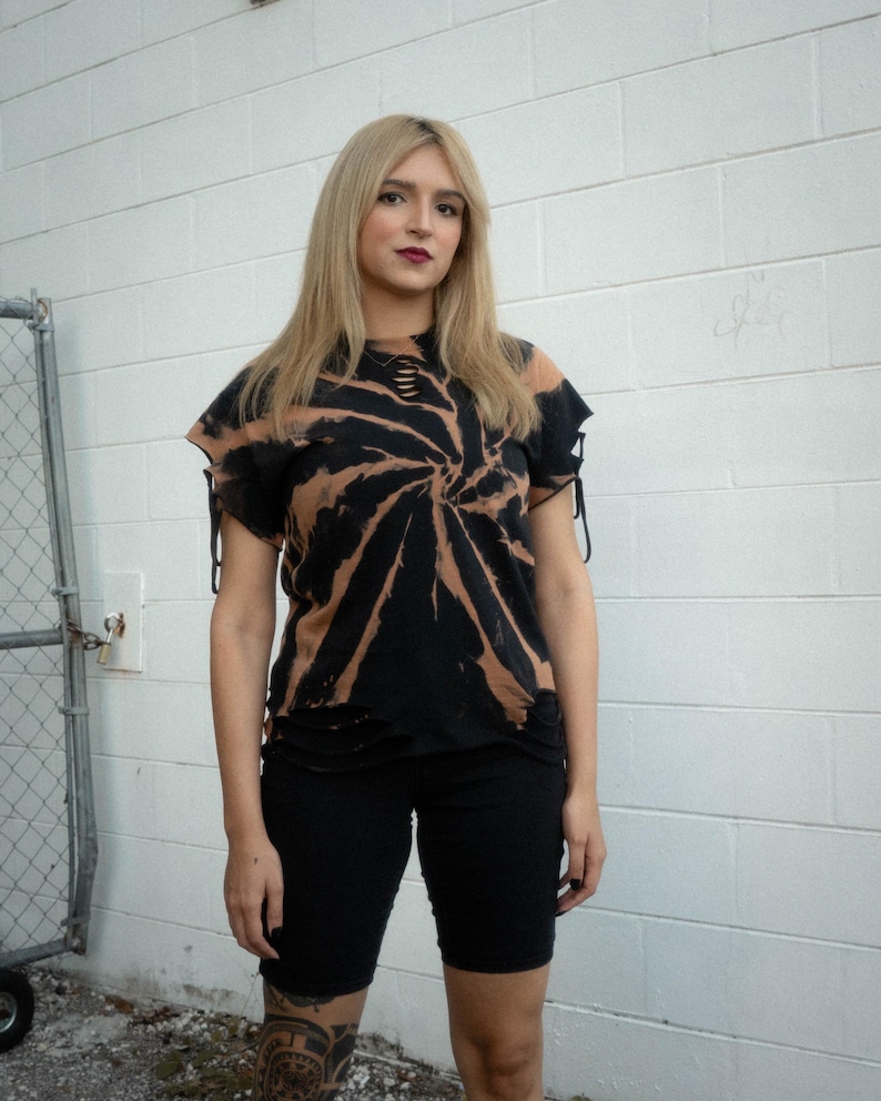 May include: A black and tan spiral tie-dye t-shirt featuring distressed cutouts at the neckline and hem, along with side sleeve ties. The shirt is paired with solid black fitted shorts that reach above the knee, creating a coordinated outfit.