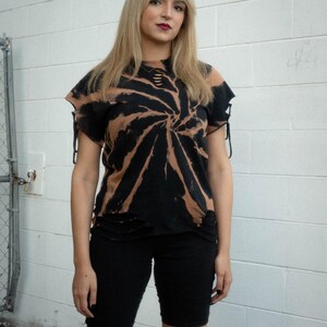 K&ouml;nnte beinhalten: Ein schwarz-beiges Batik-T-Shirt mit Spiralmuster, abgenutzten Ausschnitten am Halsausschnitt und kurzen &Auml;rmeln mit seitlichen Bindeb&auml;ndern. Das T-Shirt hat einen ausgefransten Saum und wird mit einer schlichten, eng anliegenden schwarzen Shorts kombiniert.