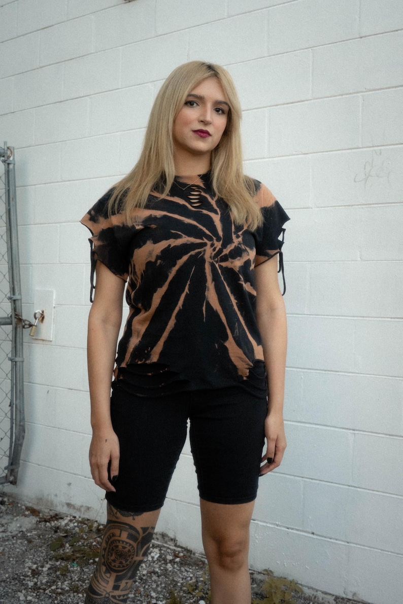 K&ouml;nnte beinhalten: Ein schwarz-beiges Batik-T-Shirt mit Spiralmuster, abgenutzten Ausschnitten am Halsausschnitt und kurzen &Auml;rmeln mit seitlichen Bindeb&auml;ndern. Das T-Shirt hat einen ausgefransten Saum und wird mit einer schlichten, eng anliegenden schwarzen Shorts kombiniert.