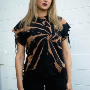 K&ouml;nnte beinhalten: Ein schwarz-beiges Batik-T-Shirt mit Spiralmuster, einem ausgefransten Ausschnitt mit horizontalen Stoffstreifen und kurzen &Auml;rmeln mit seitlicher Schn&uuml;rung. Der Saum ist ebenfalls im Used-Look mit kleinen Ausschnitten am unteren Rand gestaltet.