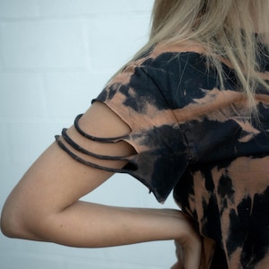 K&ouml;nnte beinhalten: Ein kurz&auml;rmeliges T-Shirt mit einem schwarz-beigen Batikmuster. Der &Auml;rmel ist mit drei horizontalen Schlitzen versehen, wodurch eine Reihe d&uuml;nner Stoffb&auml;nder entsteht, die den Oberarmbereich des Kleidungsst&uuml;cks umschlie&szlig;en.
