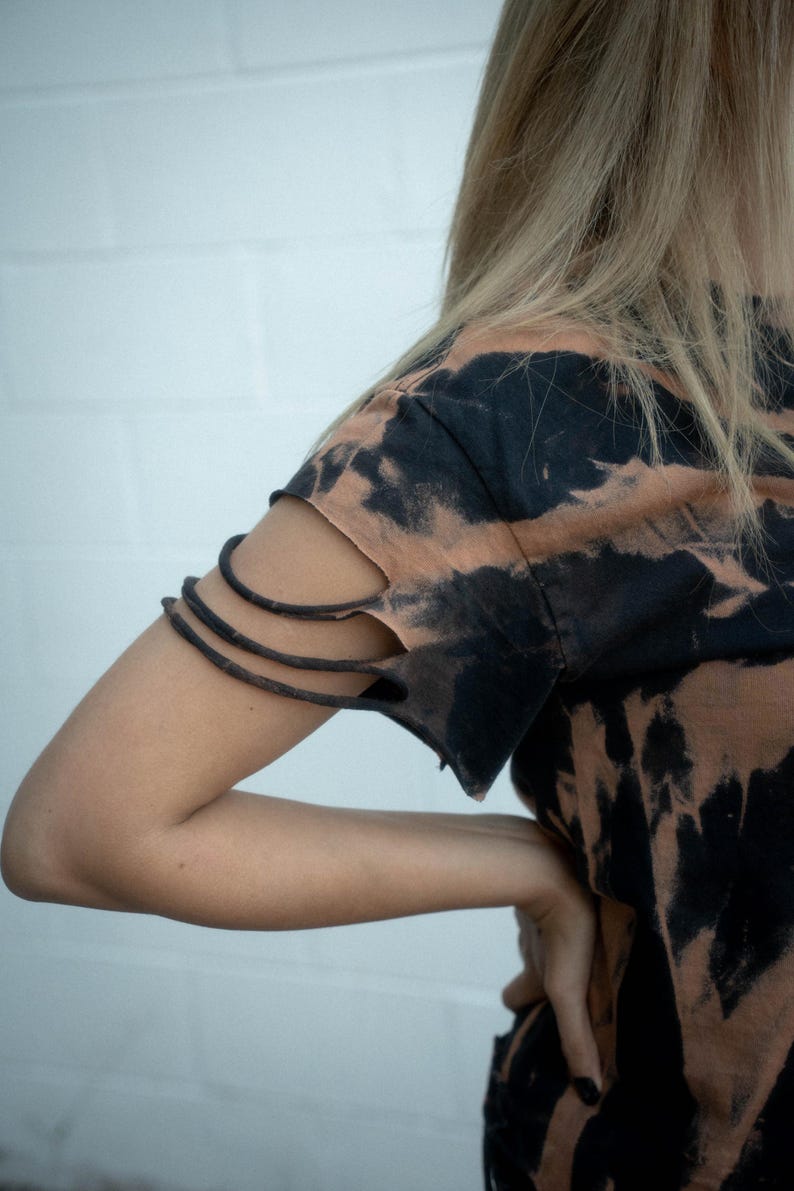 K&ouml;nnte beinhalten: Ein kurz&auml;rmeliges T-Shirt mit einem schwarz-beigen Batikmuster. Der &Auml;rmel ist mit drei horizontalen Schlitzen versehen, wodurch eine Reihe d&uuml;nner Stoffb&auml;nder entsteht, die den Oberarmbereich des Kleidungsst&uuml;cks umschlie&szlig;en.