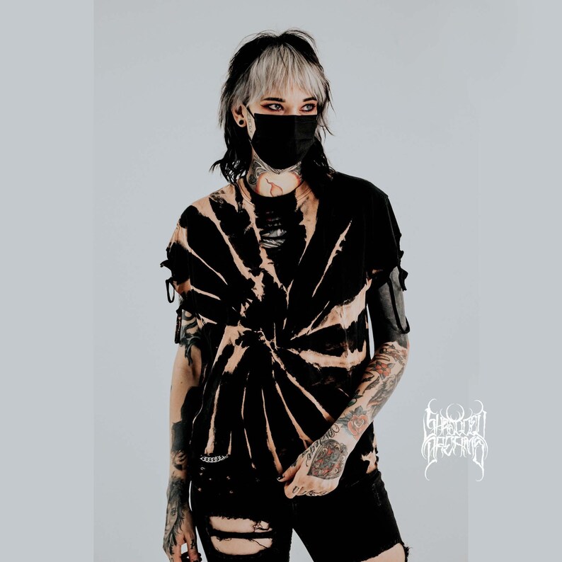 K&ouml;nnte beinhalten: Ein schwarzes T-Shirt mit gebleichtem Spiral-Tie-Dye-Muster, das an Halsausschnitt und &Auml;rmeln abgenutzte Cut-Out-Details aufweist. Das Shirt wird mit einer schwarzen, zerrissenen Jeansshorts kombiniert. Ein wei&szlig;es Logo mit stilisiertem Text ist sichtbar.