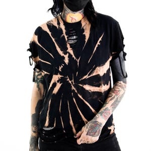 K&ouml;nnte beinhalten: Ein schwarzes Kurzarm-T-Shirt mit gebleichtem Spiral-Tie-Dye-Muster, einem abgenutzten Halsausschnitt mit horizontalen Schlitzen und seitlichen &Auml;rmeln, die mit d&uuml;nnen Stoffb&auml;ndern gerafft und ausgeschnitten sind.