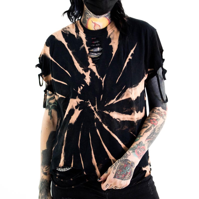 K&ouml;nnte beinhalten: Ein schwarzes Kurzarm-T-Shirt mit gebleichtem Spiral-Tie-Dye-Muster, einem abgenutzten Halsausschnitt mit horizontalen Schlitzen und seitlichen &Auml;rmeln, die mit d&uuml;nnen Stoffb&auml;ndern gerafft und ausgeschnitten sind.