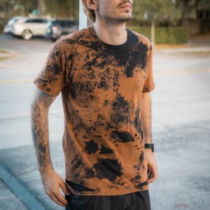 May include: A short-sleeved crew neck t-shirt featuring a distressed aesthetic with a rust-colored base and irregular black tie-dye patterns. The fabric displays small intentional holes and frayed edges throughout the garment, creating a worn look.