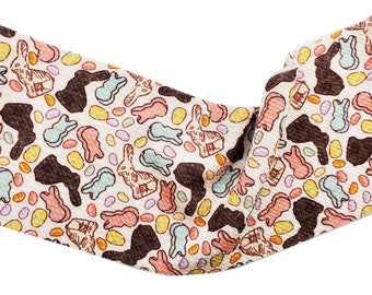 May include: A cream-colored fabric headband with a repeating pattern of Easter-themed illustrations. The design includes brown chocolate bunnies, pastel-colored marshmallow chicks, and colorful Easter eggs. The headband is twisted and knotted.
