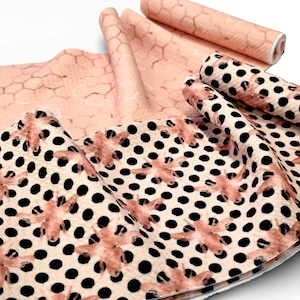 May include: Peach-coloured fabric with a honeycomb pattern and fabric with a cream background, black polka dots, and peach-coloured floral designs. The fabric is folded and rolled.