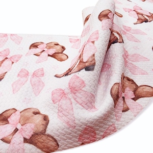 May include: White fabric with a repeating pattern of brown teddy bears and pink bows. The fabric has a textured surface and is folded, showing the pattern at different angles. The bows are a soft pink, and the bears have brown fur.
