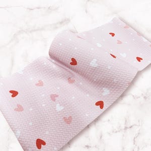 May include: Pink fabric with a textured surface, adorned with red, white, and pink heart shapes and small white dots. The fabric is folded, revealing its pattern and texture. The background is a marble pattern.
