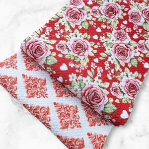 May include: Two fabric swatches. One features a red background with pink roses, white flowers, and small white hearts. The other has a white background with a repeating red floral pattern. Both fabrics have a textured surface.