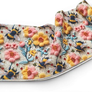 May include: A rolled textile featuring a detailed embroidered design of bees, flowers, and foliage. The flowers are in shades of yellow and pink, with blue and green leaves. The bees have black and yellow stripes. The background is a light beige.