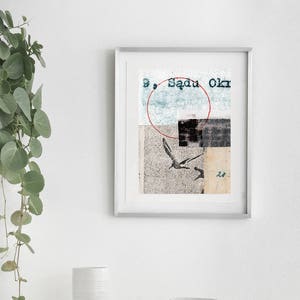 May include: A framed abstract collage print with a red circle, a bird, and the text "Sadu Okr". The print is hanging on a white wall above a white dresser with a white vase, a wooden object, and a stack of magazines.