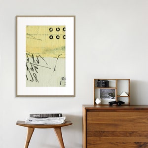 May include: A framed abstract art print with a yellow, green, and white color scheme. The print features black circles and scribbled lines. The print is hanging on a white wall above a wooden dresser and stool.