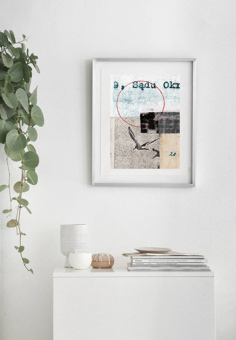 May include: A framed abstract collage print with a red circle, a bird, and the text "Sadu Okr". The print is hanging on a white wall above a white dresser with a white vase, a wooden object, and a stack of magazines.