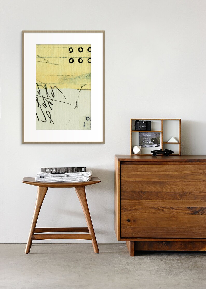 May include: A framed abstract art print with a yellow, green, and white color scheme. The print features black circles and scribbled lines. The print is hanging on a white wall above a wooden dresser and stool.