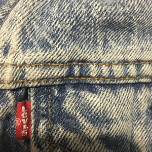 Pode incluir: Close-up de tecido de ganga azul com um remendo vermelho Levi's. O tecido tem um aspeto desbotado e desgastado e &eacute; costurado com fio amarelo.