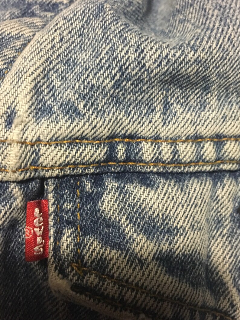 Pode incluir: Close-up de tecido de ganga azul com um remendo vermelho Levi's. O tecido tem um aspeto desbotado e desgastado e &eacute; costurado com fio amarelo.
