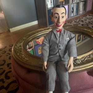 May include: A doll of a man with a large head, wearing a gray suit with a red bow tie and white shoes. The doll is sitting on a pink, tufted ottoman in front of a gold-framed mirror.