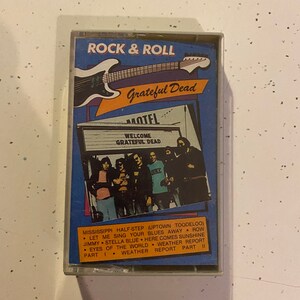 May include: A blue and yellow cassette tape with a picture of a band on the front. The band is called "Grateful Dead" and the tape is titled "Rock & Roll". The cassette tape has a list of songs on the back: "Mississippi Half-Step (Uptown Toodeloo)", "Let Me Sing Your Blues Away", "Row Jimmy", "Stella Blue", "Here Comes Sunshine", "Eyes of the World", "Weather Report Part I", "Weather Report Part II".
