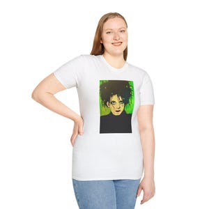 May include: White t-shirt featuring a portrait of a person with dark, voluminous hair and a black turtleneck. The portrait has a green and yellow background. The person is wearing blue jeans.