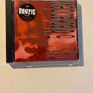 May include: A CD case featuring the album "Danzig 6:66 Satan's Child." The cover is predominantly red with vertical white lines and the band's name in large, stylized text. A black circle indicates "NEW" and "featuring sacrifice."