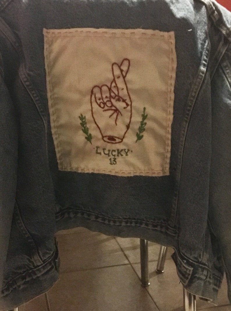 Pode incluir: Uma jaqueta de ganga azul com um remendo bege costurado nas costas. O remendo apresenta uma imagem bordada de uma m&atilde;o com os dedos cruzados, dois ramos verdes e o texto "LUCKY 13".