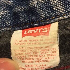 Pode incluir: Um r&oacute;tulo de cuidados Levi's branco com texto vermelho. O r&oacute;tulo tem instru&ccedil;&otilde;es para lavar e secar a roupa. O texto diz: "Para garantir um ajuste adequado, experimente esta roupa antes de comprar. Lavar na m&aacute;quina com &aacute;gua quente. Ciclo normal. Secar na m&aacute;quina a baixa temperatura. Lavar e secar com cores semelhantes. A cor pode transferir ao lavar pela primeira vez. Lavar antes de usar. Feito nos EUA. Conte&uacute;do no verso."